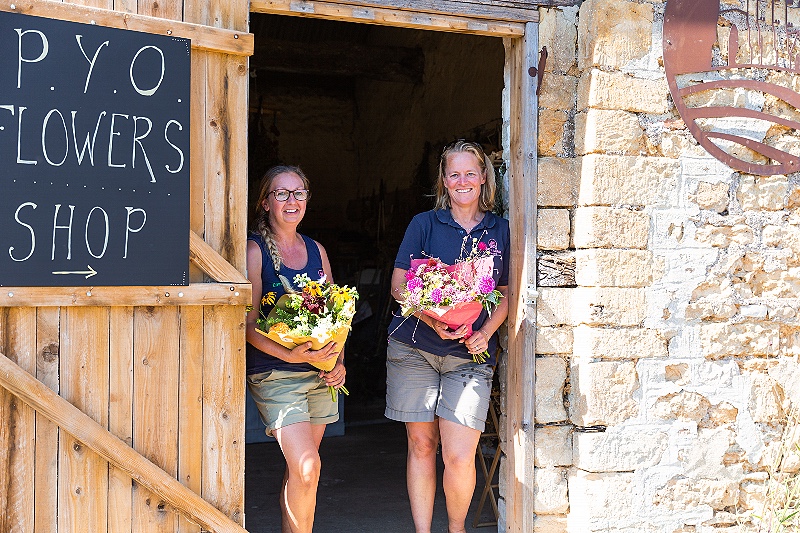 Flower farmers holding wrapped bouquets of freshly cut seasonal flowers stand in the doorway of a stone barn waiting to welcome visitors to their pick your own session as part of The Flower Farmers' Big Weekend.