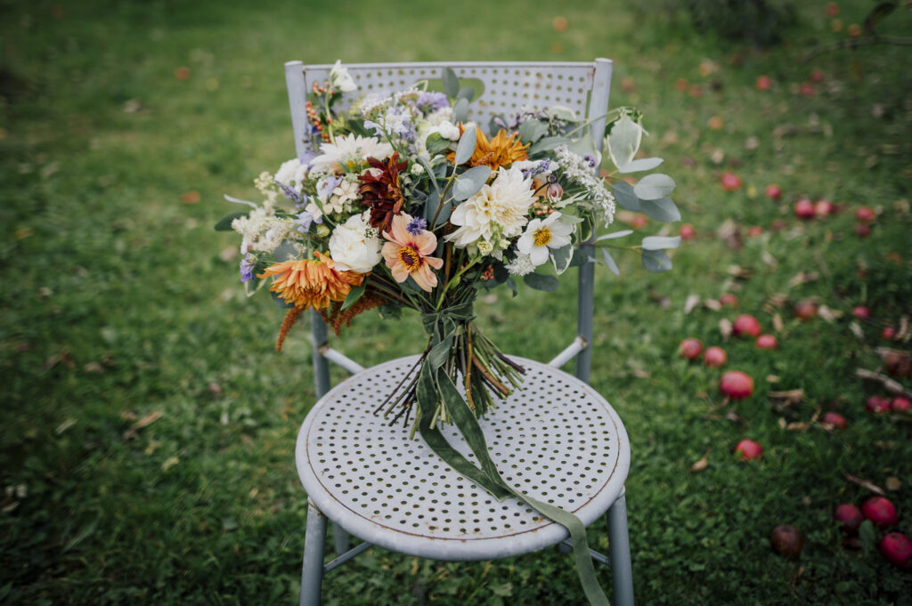 A pastel bouquet with pops of orange by Camomile and Cornflowers stands on a metal chair on the grass. Photo by Dearest Love Photography.