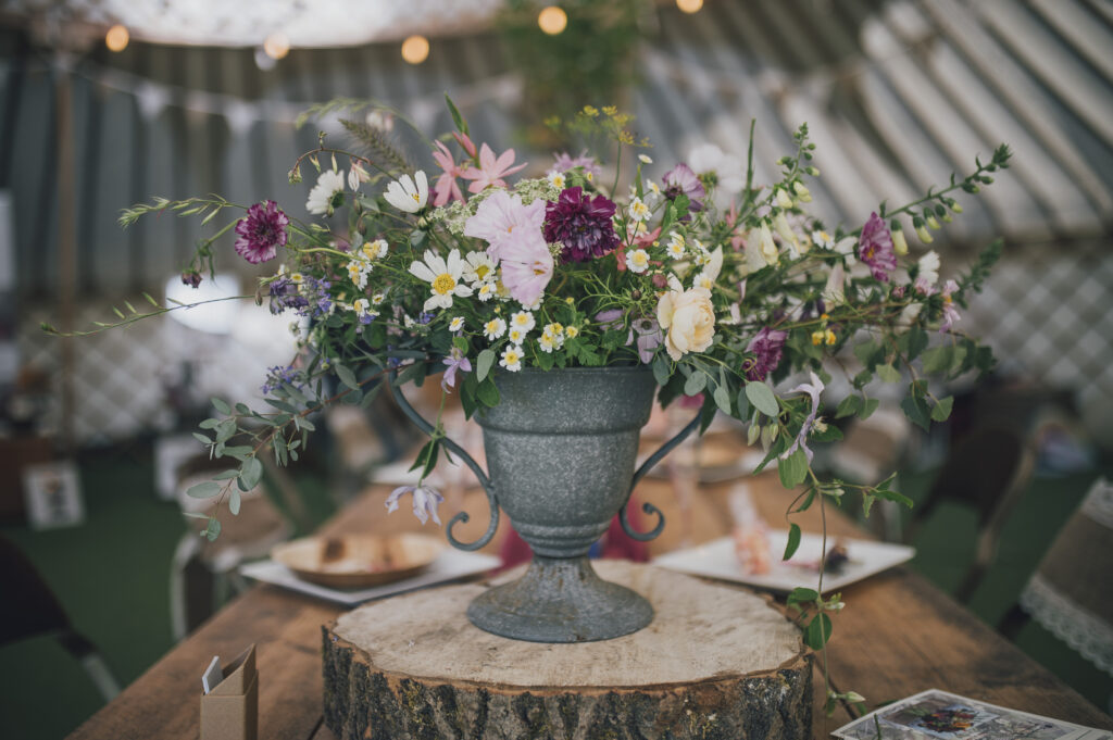 A wedding urn filled with pastel summer flowers stands on a log slice in a marquee. Camomile and Cornflowers. Photo by Dearest Love Photography.