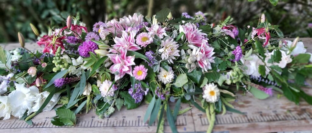 A pink summer coffin spray by Camomile and Cornflowers rests on a table.
