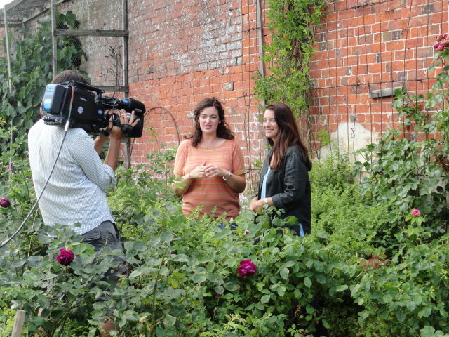 Rachel de Thame discusses cut flower growing for the British Garden Revival programme with Catkin's Rachel Petheram in the walled garden at Doddington Hall. A cameraman films them chatting in front of a beautiful brick wall with old rambling roses scrambling up it.