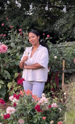 Sophia is pictured outdoors standing next to a bed of flourishing summer dahlias holding a glass of champagne in celebration.