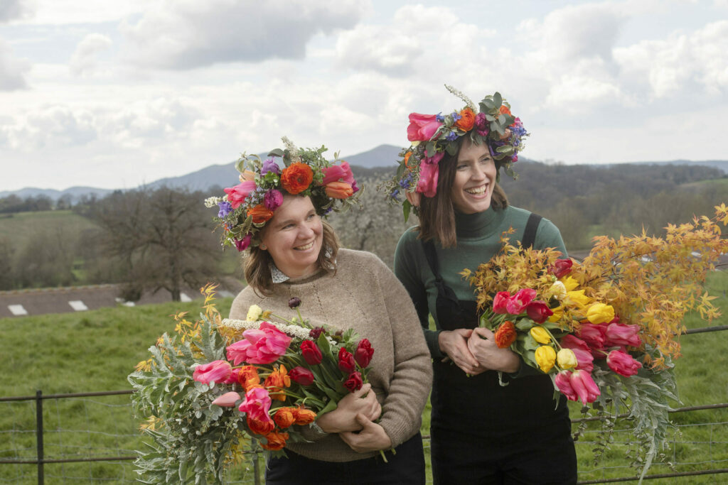 Hannah Walker and Meg Edmonds pictured at Meg's farm with the Malvern Hills in the background wearing brightly coloured flower crowns holding armfuls of late Spring flowers