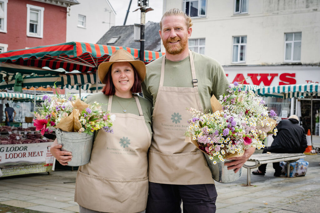 Clare and Simon of Blossoms and Berries holding buckets of their fresh cut flowers at a local farmers' market in Pembrokeshire