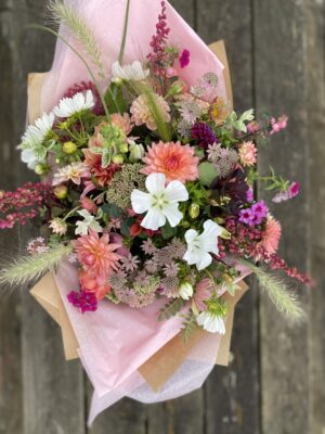 A peachy late summer bouquet with dahlias, grasses, white malope and cosmos daisies wrapped in paper and sitting on a wooden surface.