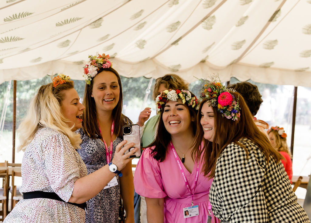 A group of women laugh as they pose for selfies wearing fresh flower crowns in the flower school marquee at RHS Hampton Court 2023