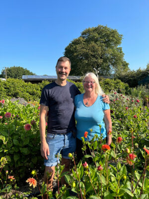 Nicolaus Peters and Carol Siddorn stand in Carol's dahlia plot surrounded by scarlet blooms and the Cheshire countryside forming a sunny summer backdrop.