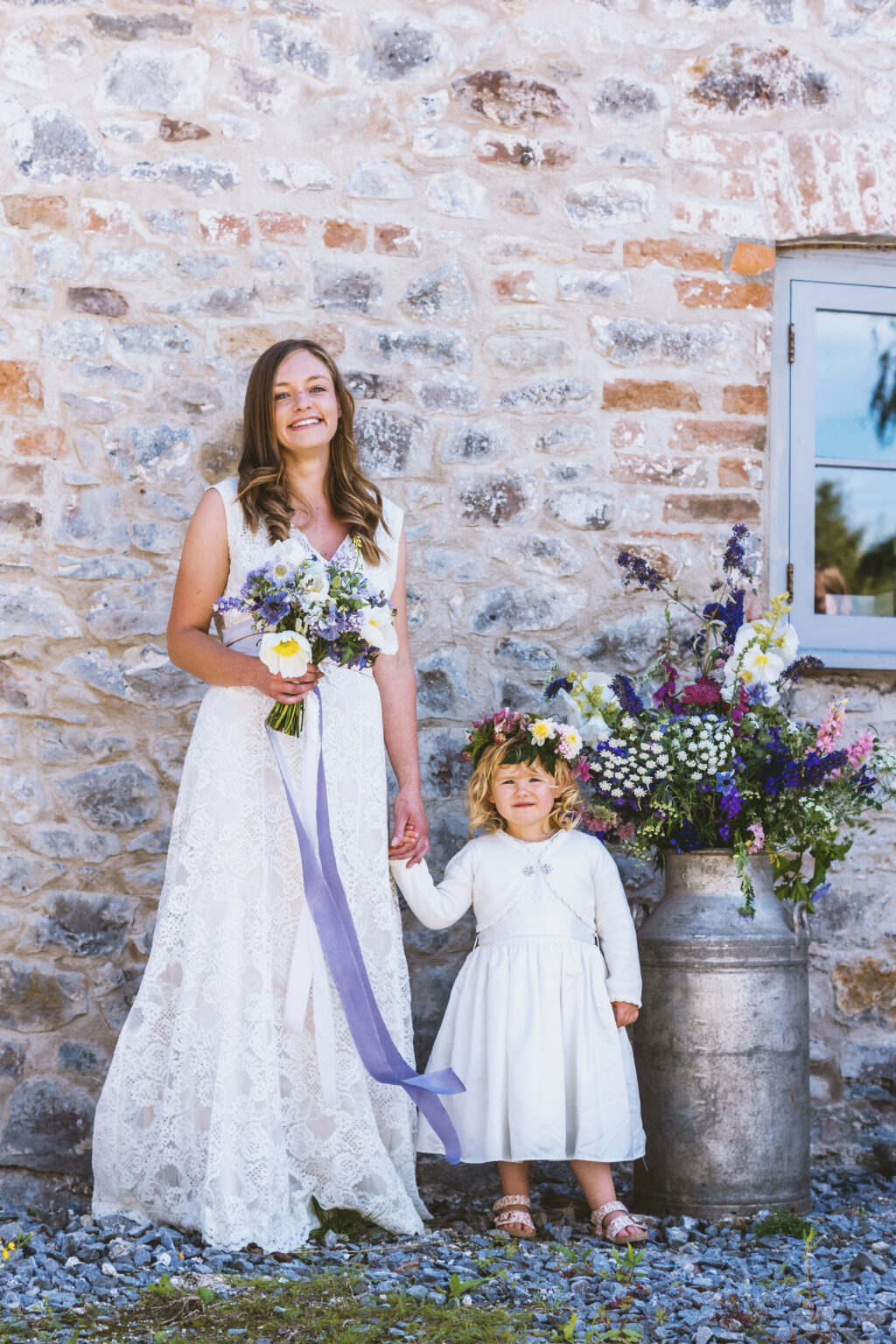 A bride and flower girl with British grown wedding flowers by The Floral Potager stand smiling against a rustic brick wall