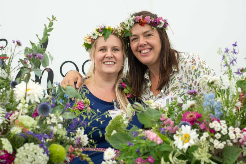 Claire and Alison of The Flower Farm pose in flower crowns surrounded by a sea of summer flowers.