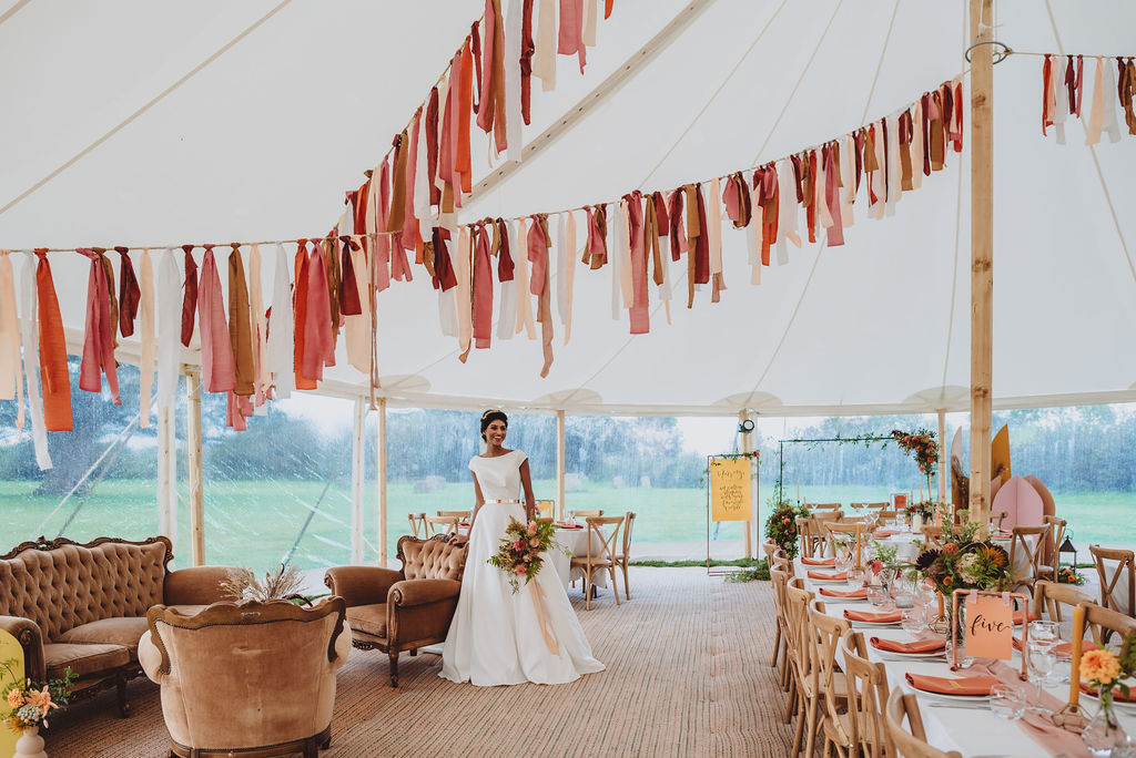 A bride stands with her late summer bouquet in a tipi wedding decorated with bunting overhead and dahlias on the long trestle tables for guests