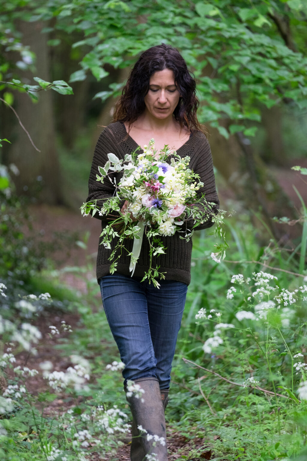 Emma de Sousa, Urban Flower Farmer walks along a grassy path, surrounded by hedges and greenery, holding a seasonal wedding bouquet of flowers she has grown in her cutting garden. Photo: Emma Davies Photography.