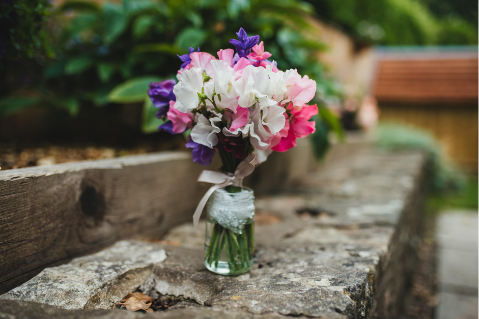 A posy of freshly cut sweet peas in shades of pink, white and purple, tied with a simple ribbon stand in a jar on top of a rough stone wall. Photo: Compton Garden Flowers.