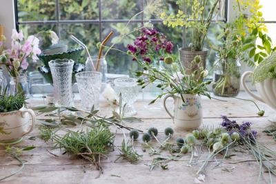 wedding flower preparations in progress. stems of chinos, lavender and herbs lie on a wooden worktop which also holds pretty vintage jugs filled with garden gathered cut flowers.