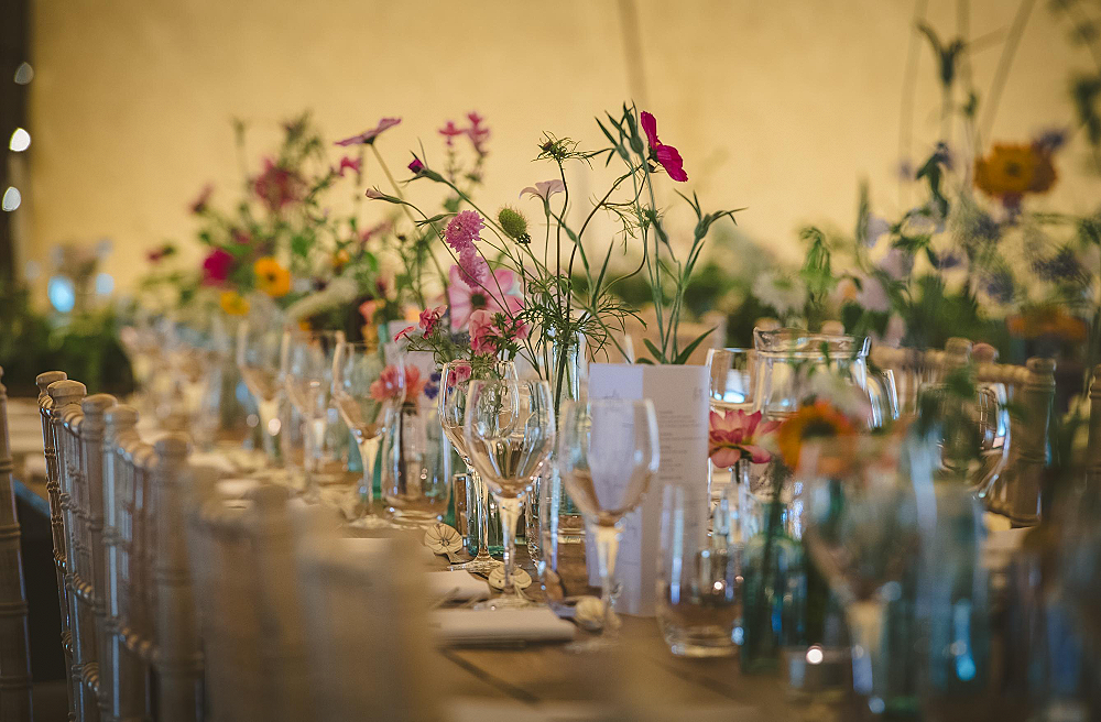 Bright July flowers in vintage bottles decorate a rustic wooden guest table at an outdoor tipi reception.