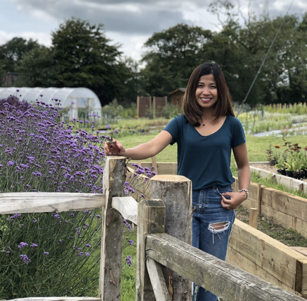 Aizel Finch of Yalham Hayes Flower Farm stands smiling by a fence on her flower plot with purple verbena bonariensis billowing out of the borders in high summer.